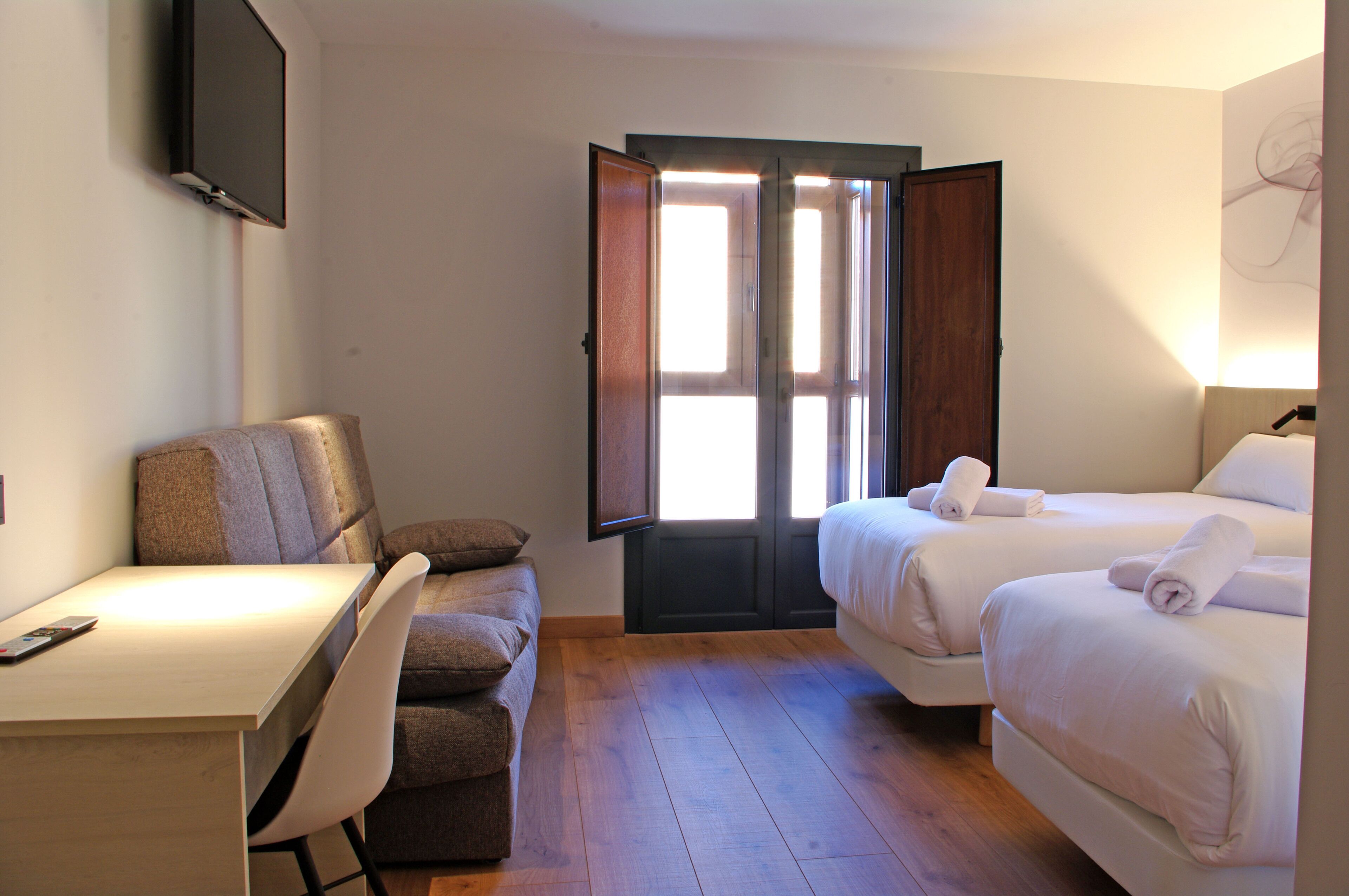 Habitación de hotel con escritorio, dos camas, una silla y un ventanal grande.