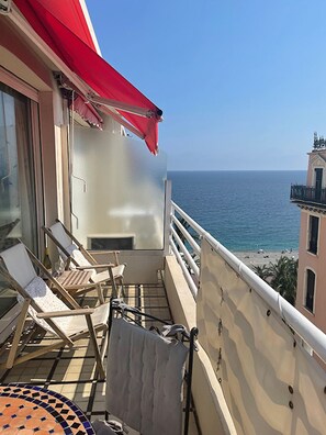 A balcony with two lounge chairs, a small table, and a red awning. The balcony overlooks a beach and the sea.