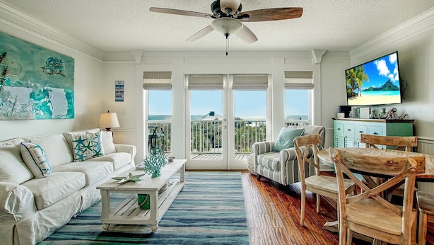 Uma sala de estar com ventilador de teto, uma janela grande com vista para o mar, uma televisão e uma mesa de centro de madeira.