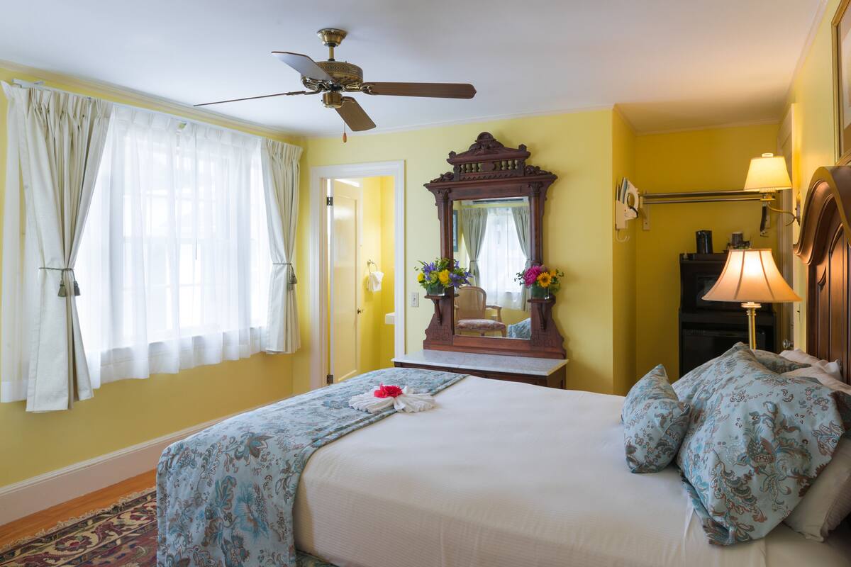Una habitación de hotel con cama, ventilador de techo, espejo, lámpara y ventana con cortinas.