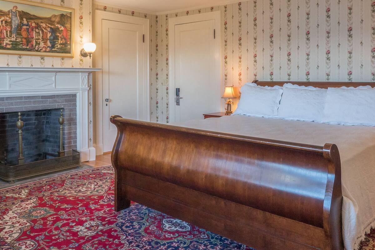 Un dormitorio tradicional con cama de madera, chimenea, papel tapiz floral y alfombra estampada.
