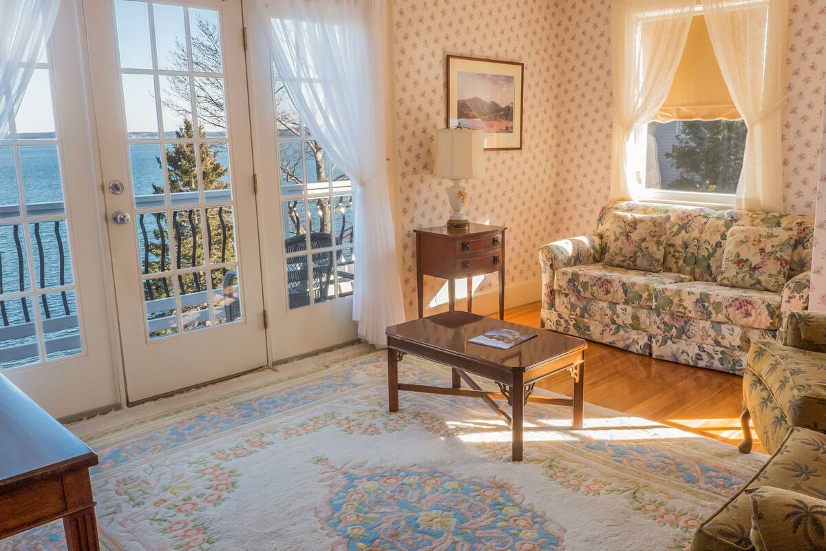 Una sala de estar con sofá, sillón y una mesa. Hay una ventana con cortinas, un cuadro en la pared y vista al mar a través de puertas de cristal.