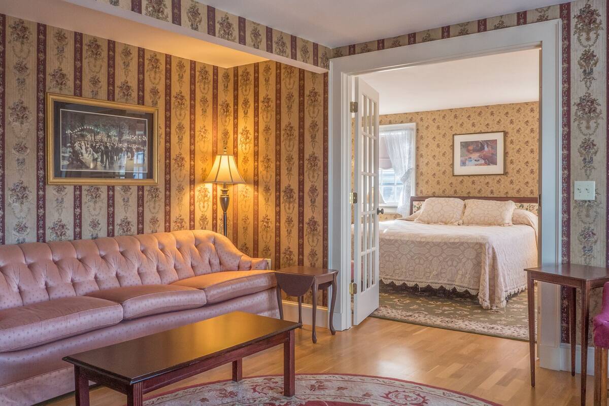 Una habitación con un sofá rosa, una mesa de centro de madera, un sillón morado, una cama con colcha blanca y una lámpara.