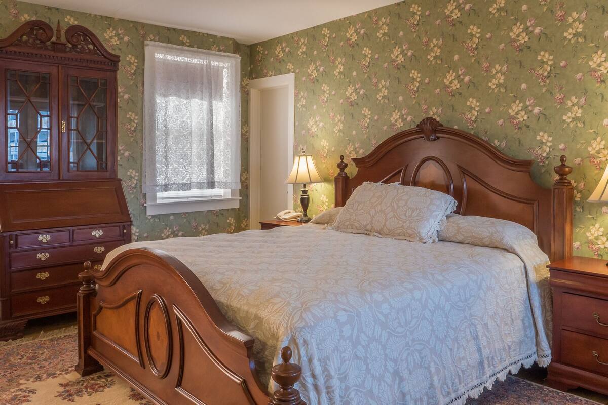Un dormitorio con una cama de madera, una mesita de noche, un aparador y una ventana con cortina de encaje.