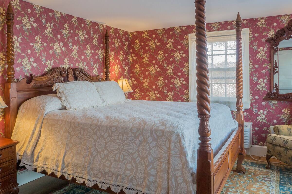 Una cama con dosel con papel tapiz floral, una mesita de noche de madera con lámpara y un sillón con cojín estampado.