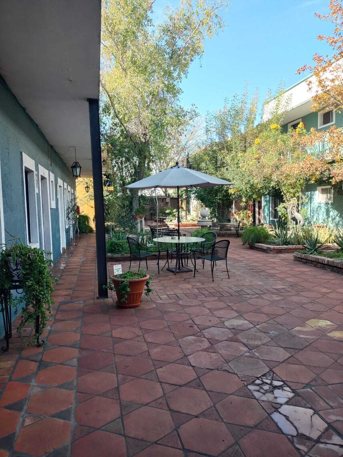An outdoor patio area with a table and chairs under a large umbrella, surrounded by potted plants and a tiled floor.