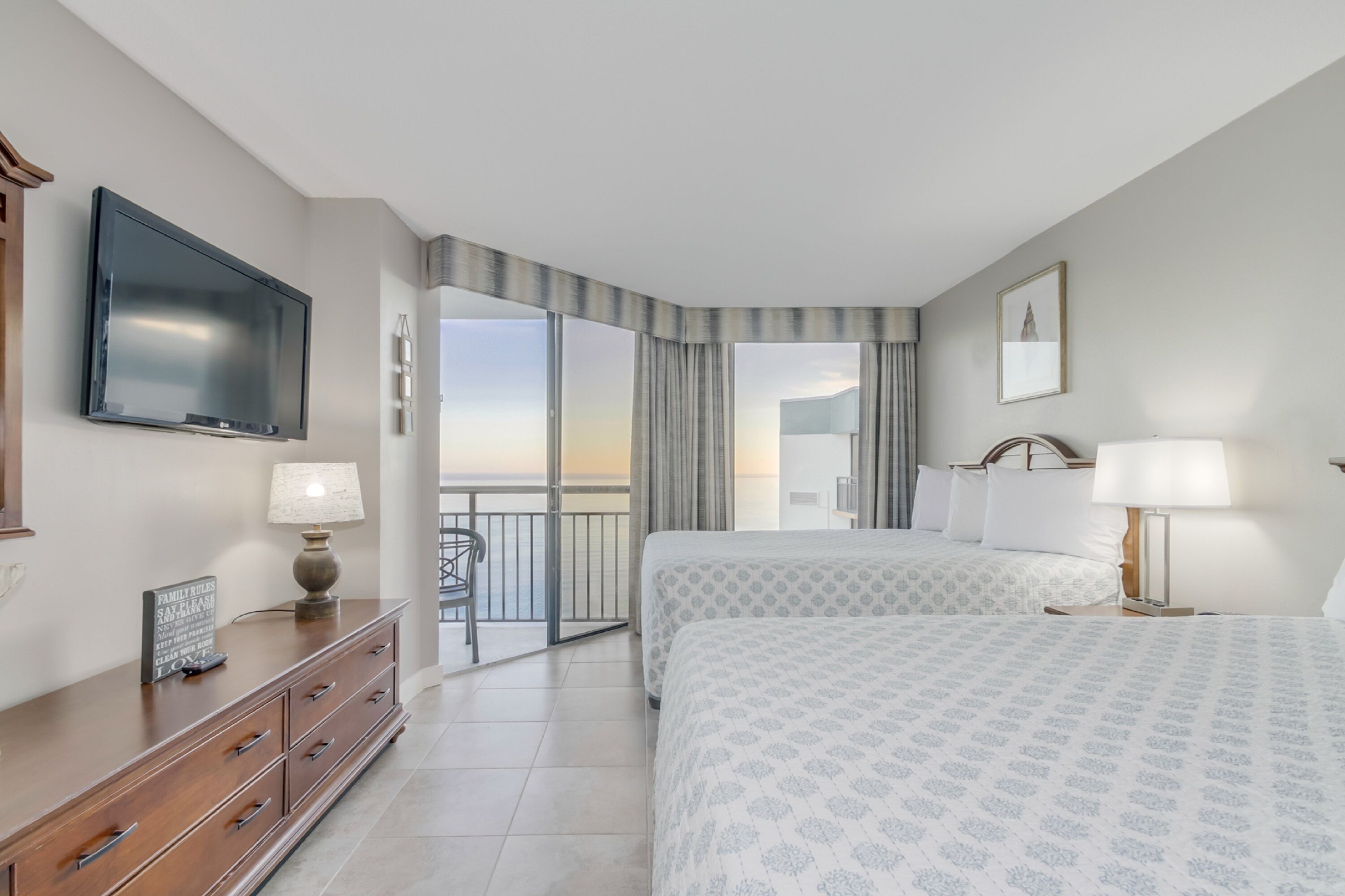 Een hotelkamer met een groot bed, een aan de muur bevestigde televisie, een dressoir en een balkon met gordijnen.