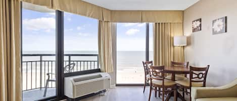 Une chambre d’hôtel avec un balcon donnant sur la plage, un coin repas avec des chaises en bois et une grande fenêtre avec des rideaux.
