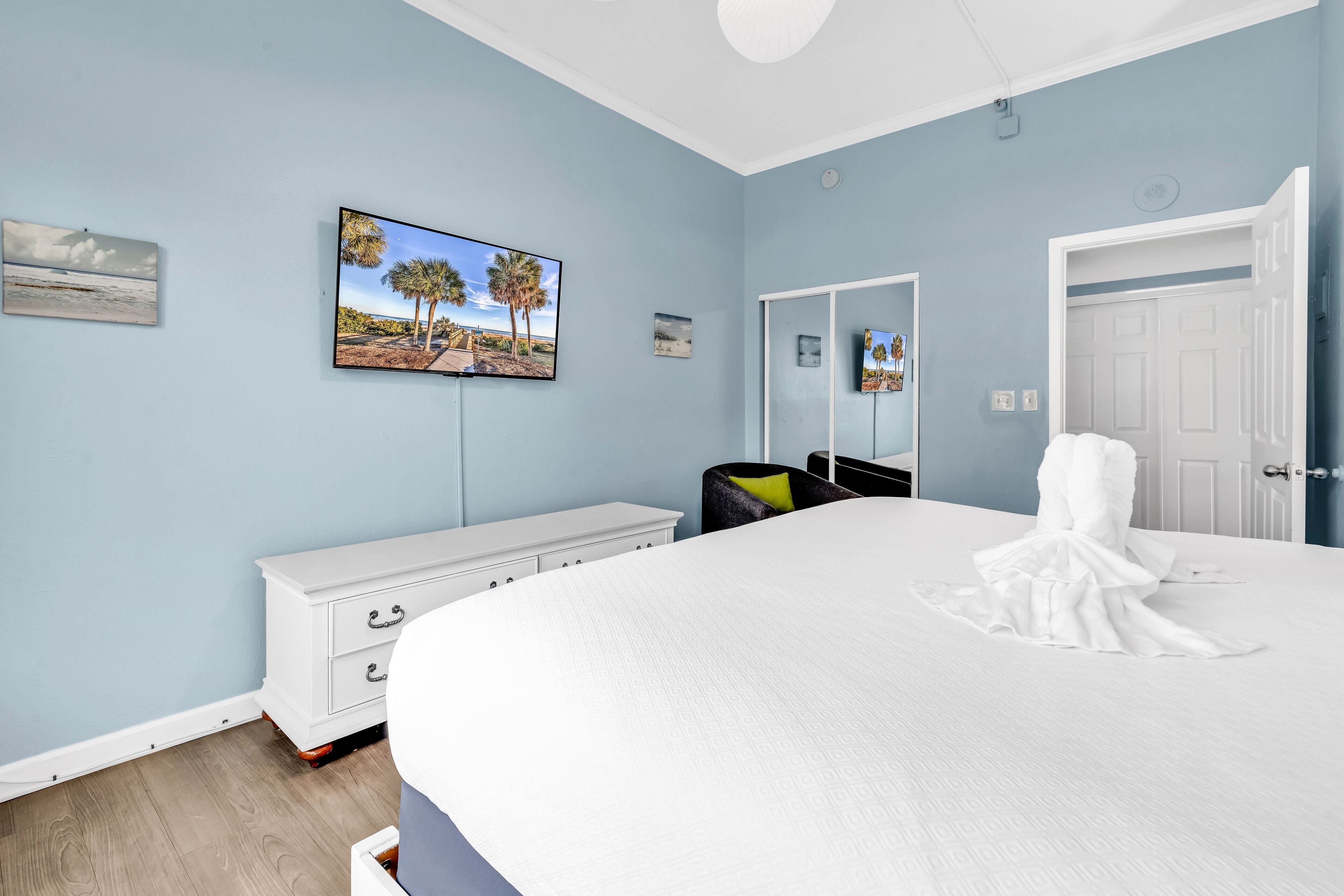 Une chambre à coucher avec un grand lit, une commode blanche, un téléviseur fixé au mur et une porte.