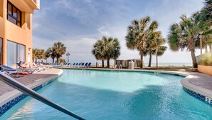 A swimming pool with lounge chairs and palm trees, surrounded by a building with a balcony. - Ocean Front Condos at Patricia Grand (Myrtle Beach)