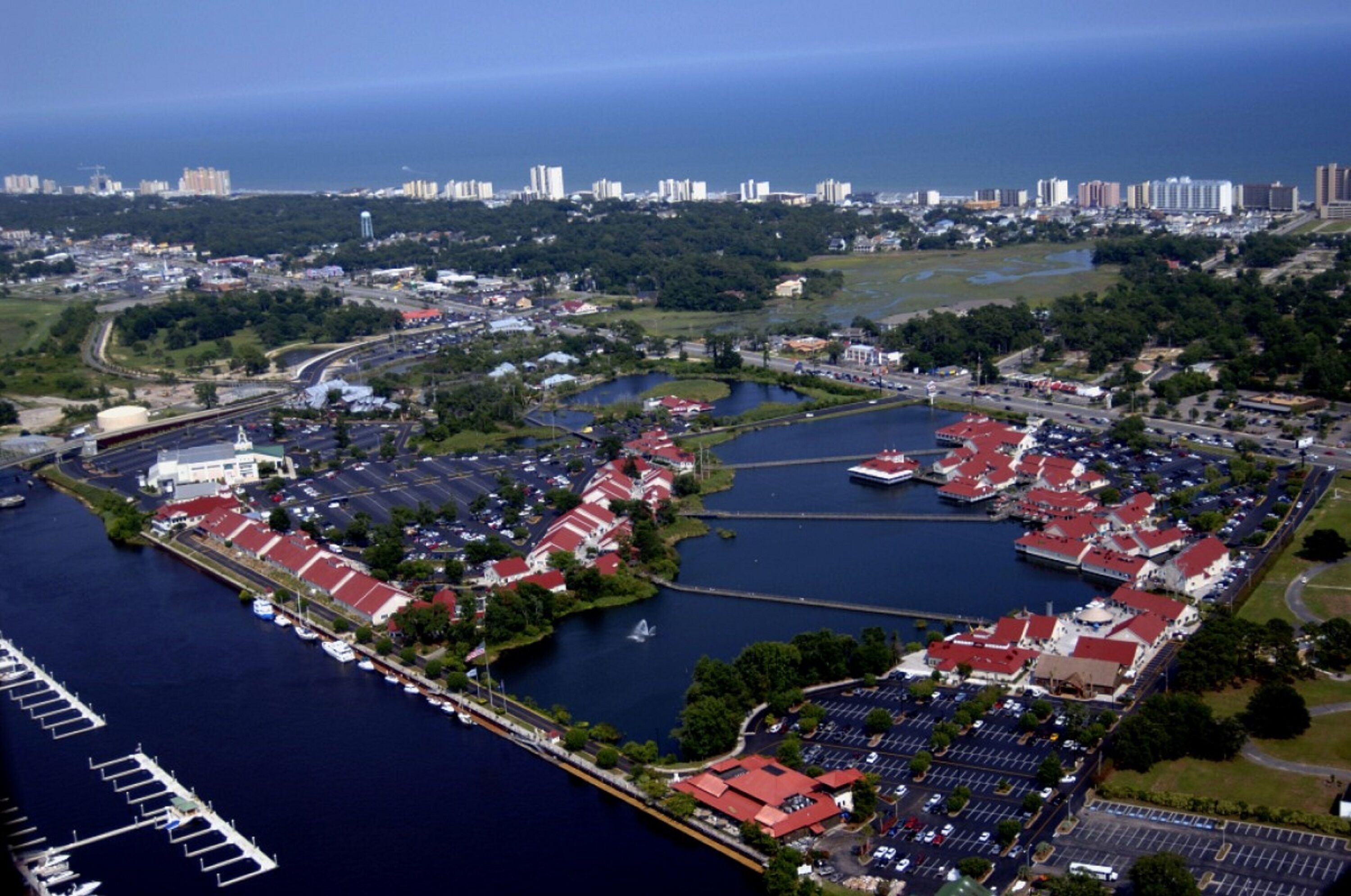 An aerial view of a marina with boats docked, a large parking area, and a city skyline in the background.