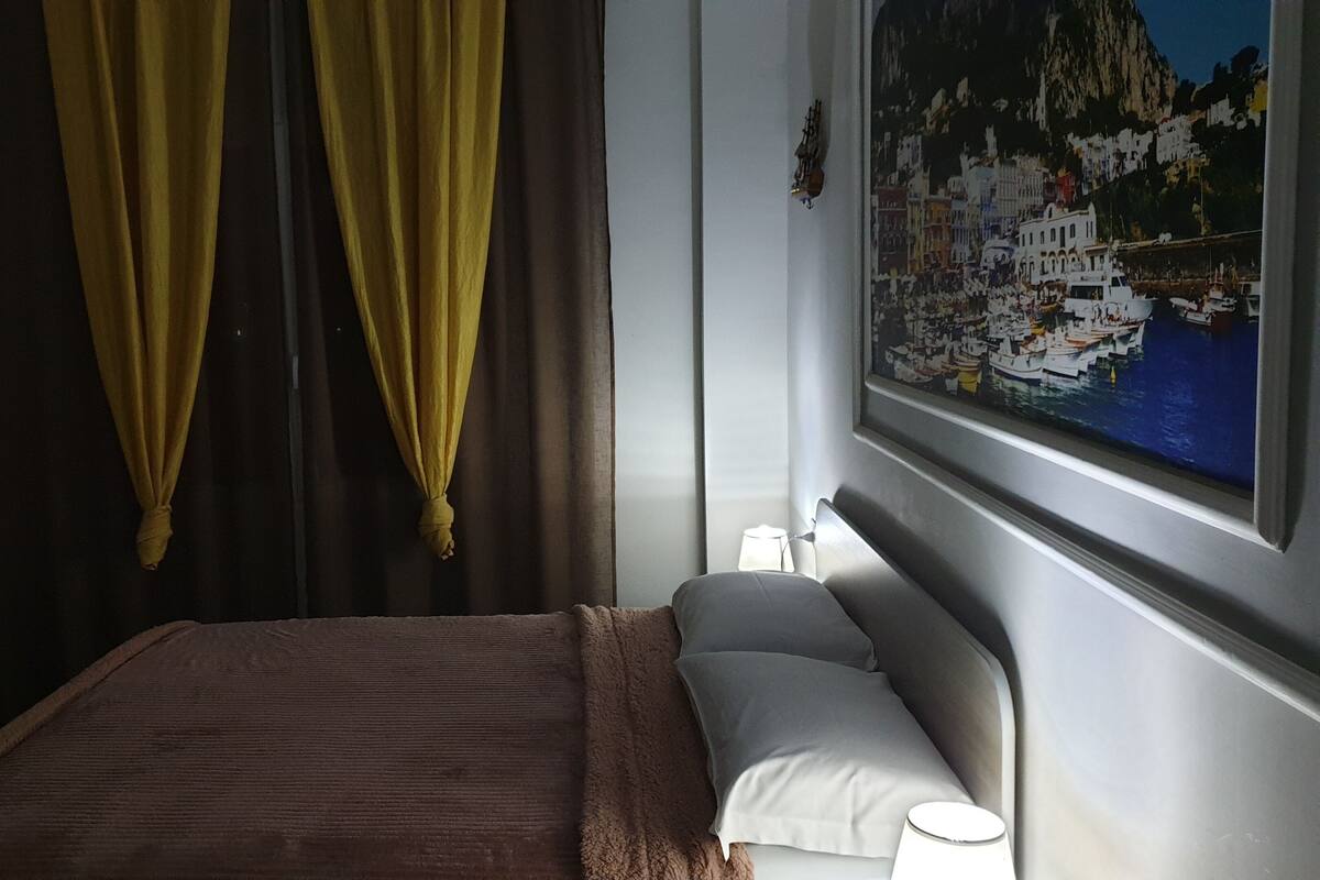 Een hotelkamer met een bed, nachtkastje, lamp en een schilderij van een kustplaats.