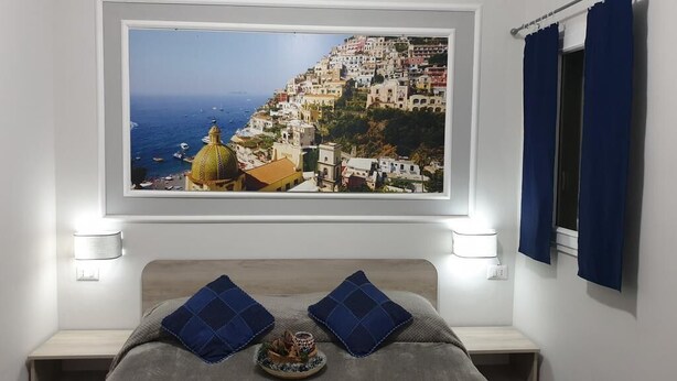 A bedroom with a large window overlooking a coastal town, a bed with blue pillows, and bedside lamps. - Oh Mamma Mia (Pompei)