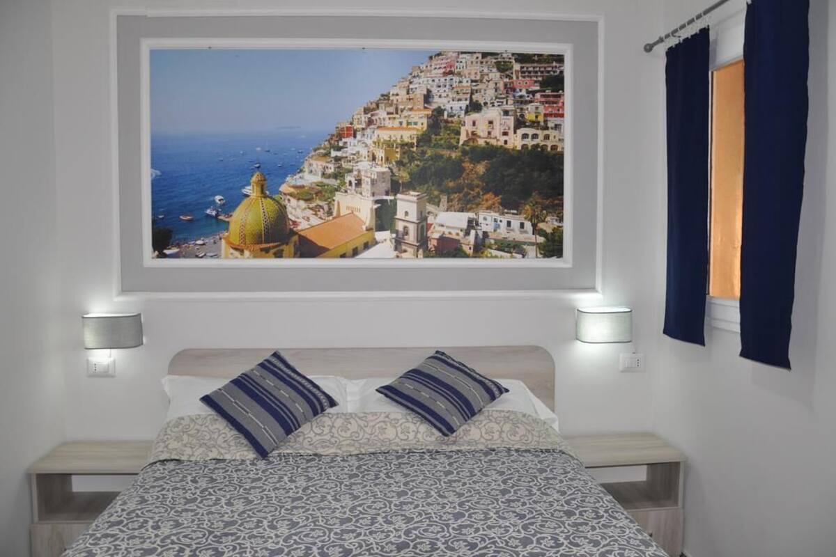 Een slaapkamer met een bed, twee bedlampen, een raam met uitzicht op een kustplaats en een gordijn.