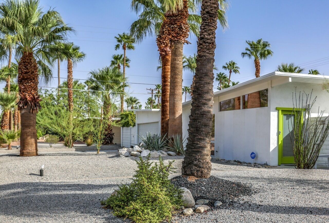 A modern building with a green door, surrounded by palm trees and a gravel driveway.