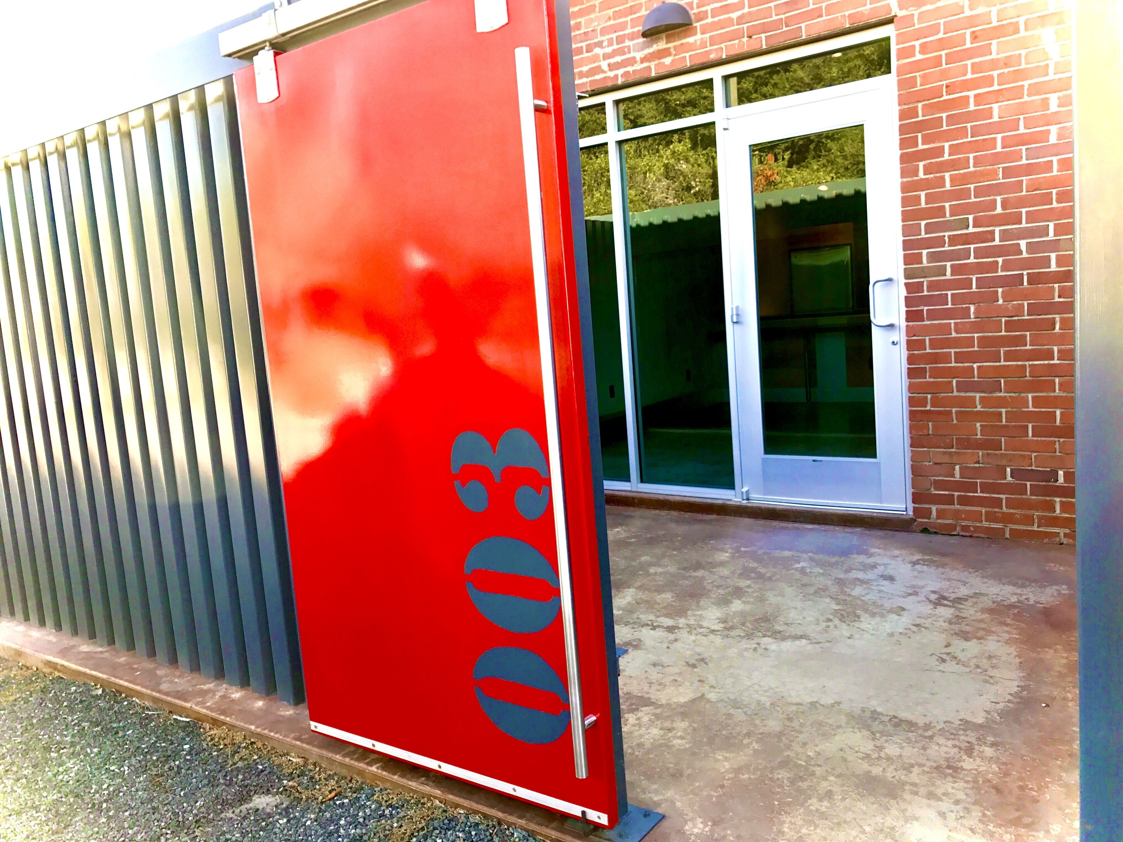 Una puerta metálica roja con el número 303, una puerta de vidrio con marco blanco, y un edificio de ladrillo al fondo.