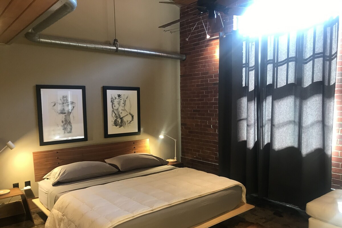 A bedroom with a bed, two framed artworks, a wooden headboard, a nightstand, a bedside table, and a window with a curtain.