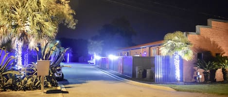 Straßenansicht bei Nacht mit einem Gebäude auf der rechten Seite, Palmen und dekorativer Beleuchtung.