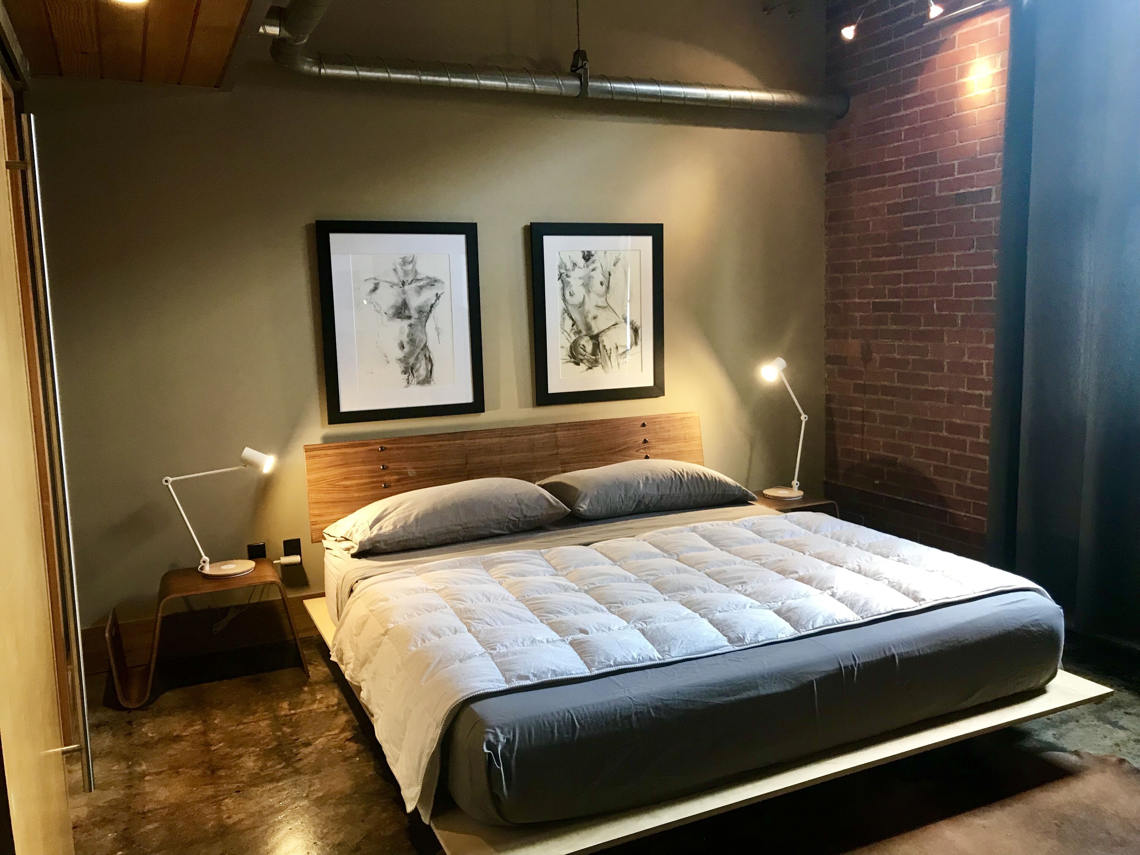A modern bedroom with a wooden headboard, a large bed with a quilted white duvet, two framed artworks on the wall, and a nightstand with a lamp.