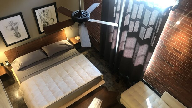 A bedroom with a bed, a ceiling fan, a nightstand, a lamp, a window with curtains, and a brick wall. - The Hangar Lofts Hotel (Columbia)