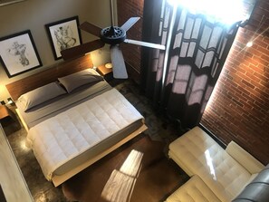 A bedroom with a bed, a ceiling fan, a nightstand, a lamp, a window with curtains, and a brick wall. - The Hangar Lofts Hotel (Columbia)