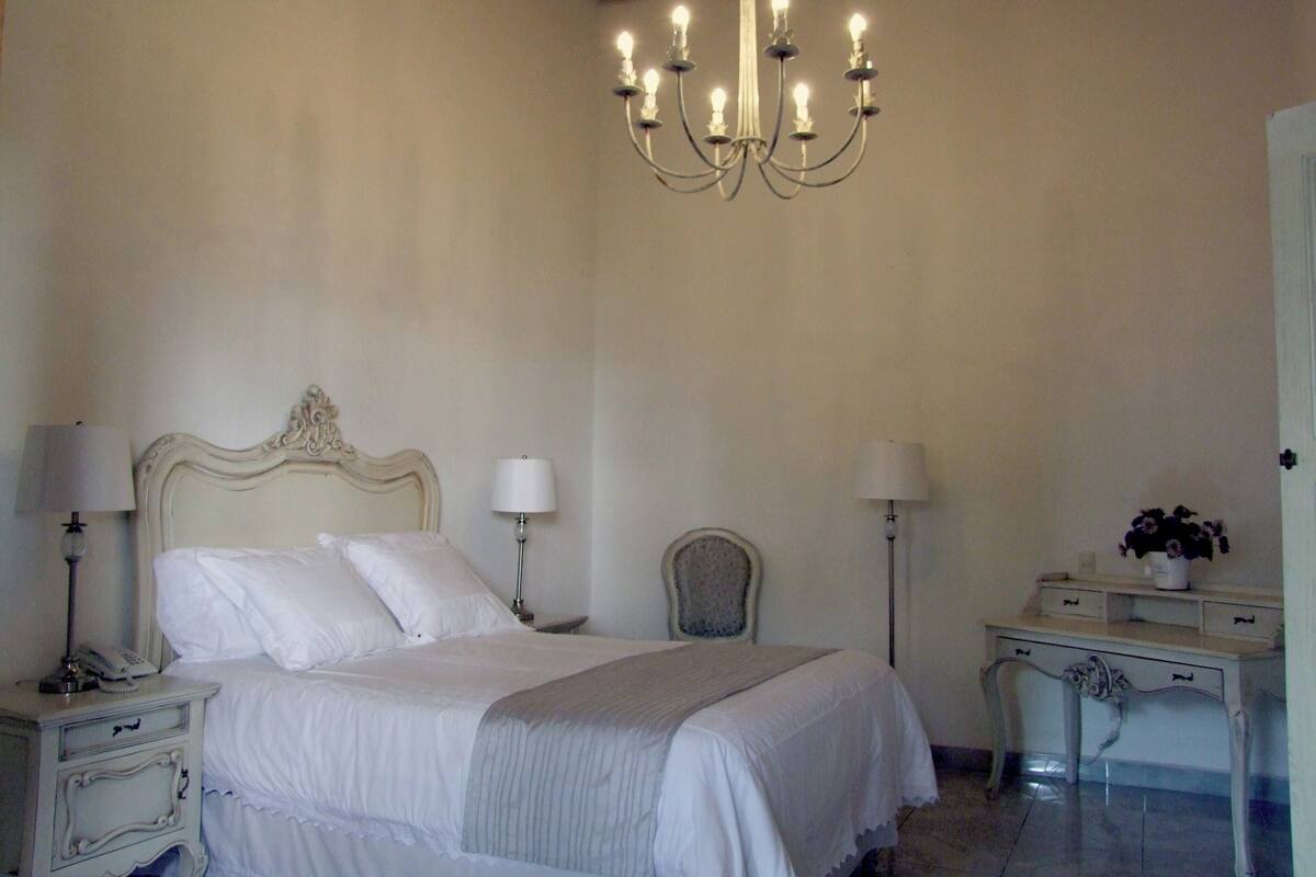 A bedroom with a large bed, two bedside tables, a chair, a desk, and a chandelier.