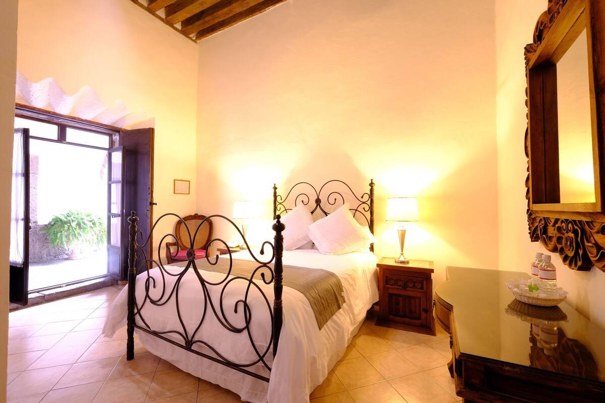A bedroom with a bed, a nightstand, a table with a lamp, and a mirror.