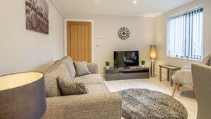 A modern living room with a sofa, a TV, a lamp, and a window with blinds. - 14 St Johns Mews - sleeps 3 guests  in 1 bedroom (York)