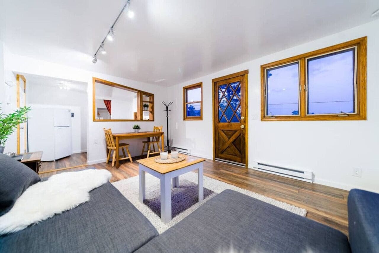 Una sala de estar moderna con un sofá, una mesa de centro y un comedor con mesa y sillas. Se ve una ventana grande con vistas al cielo.
