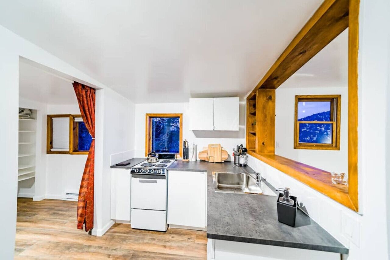 Una cocina compacta con gabinetes blancos, estufa y pileta. Se observan vigas de madera en el techo y una ventana con vista a la montaña.
