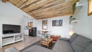 Uma sala de estar moderna com teto de madeira, uma TV de tela plana, um sofá, uma mesa de centro e um espaço de jantar com cadeiras e uma mesa.