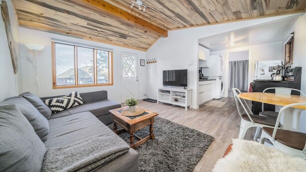 Uma sala de estar moderna com um sofá cinzento, mesa de centro de madeira e uma televisão. O espaço possui teto de madeira e uma área de cozinha com mesa e cadeiras.