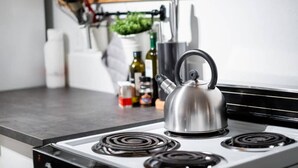 Uma cozinha com uma chaleira de aço inoxidável sobre o fogão, uma estante de madeira com utensílios de cozinha e vários frascos e recipientes na bancada.