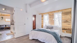 Quarto com cama, cabeceira de madeira, porta branca e vista para uma sala de estar.