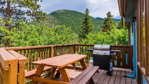 Um deck de madeira com mesa de piquenique e churrasqueira, com vista para uma montanha arborizada.