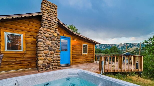 Een houten chalet met een stenen schoorsteen, een hottub en een terras met uitzicht op een bergachtig landschap.
