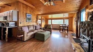 Een knus interieur van een chalet met houten plafond, een open haard, een eethoek en een zithoek met een bank en een poef.