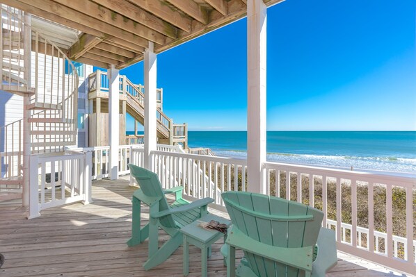 A wooden deck with chairs and a table, overlooking a beach with a clear view of the ocean.