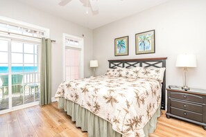A bedroom with a bed, a nightstand, a dresser, a window with a view of the ocean, and two framed pictures on the wall.