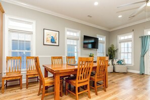 A dining area with a wooden table and chairs, a flat-screen TV mounted on the wall, and large windows with blinds. - Anchors Away - Stunning 5BR Oceanfront Home - Elevator, Comm. Pool (Surf City)