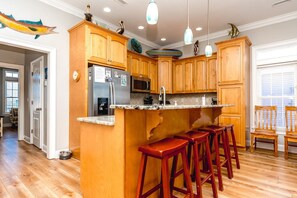 A kitchen with wooden cabinets, a stainless steel refrigerator, a microwave, and a granite countertop. There are four red bar stools and a window with blinds. - Anchors Away - Stunning 5BR Oceanfront Home - Elevator, Comm. Pool (Surf City)