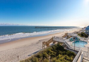 A beach with a wooden pier extending into the sea, a pool area with a deck, and a clear sky.