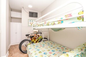 A bunk bed with a floral-patterned mattress, a bicycle, a fan, and a white wardrobe in a room with white walls and a ceiling light.