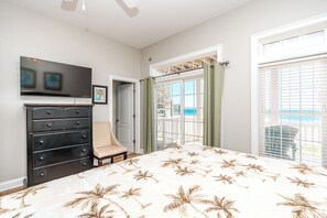 A bedroom with a bed, a dresser, a chair, a television, and a door leading to a balcony with a view of the beach.