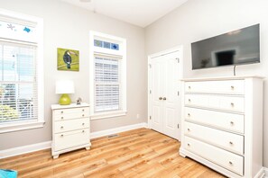 A room with a wooden floor, white furniture, a television mounted on the wall, and windows with blinds.