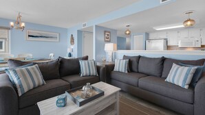A living room with two grey sofas, a coffee table with decorative items, a chandelier, and a kitchen in the background. - Starboard Village Unit 225 (Pensacola Beach)