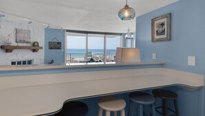 A room with a view of the ocean, a bar area with stools, and a hanging light fixture. - Starboard Village Unit 225 (Pensacola Beach)