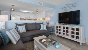 A living room with a grey sofa, striped pillows, a coffee table with decorative items, a TV on a white cabinet, and a kitchen in the background. - Starboard Village Unit 225 (Pensacola Beach)