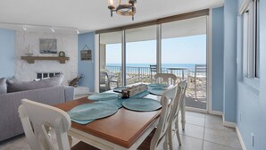 A dining area with a wooden table, white chairs, and a view of the beach through large sliding glass doors. - Starboard Village Unit 225 (Pensacola Beach)