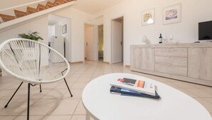 A modern kitchen with a white chair, a dining table with books, and a wooden cabinet with a TV. - Blu Mediterraneo House - 1 (Ispica)